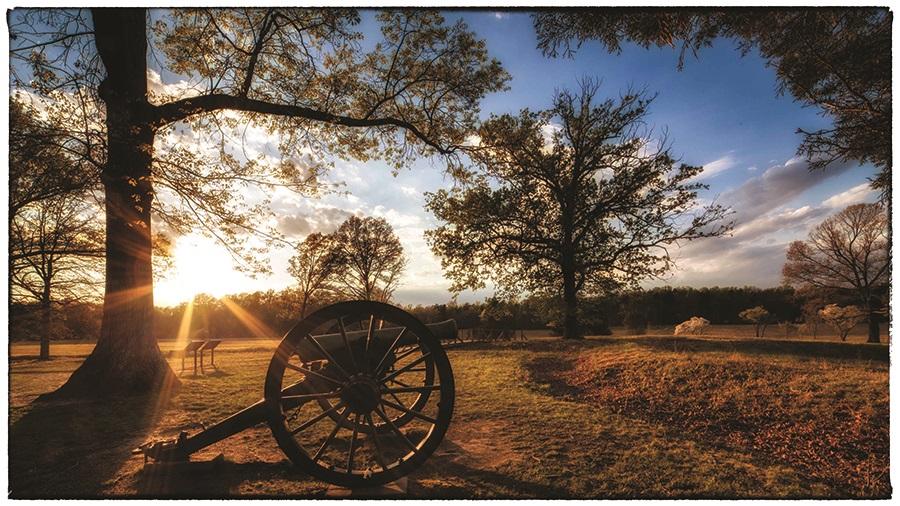 This is a photograph of a cannon with he sun shining through the trees in the background. 