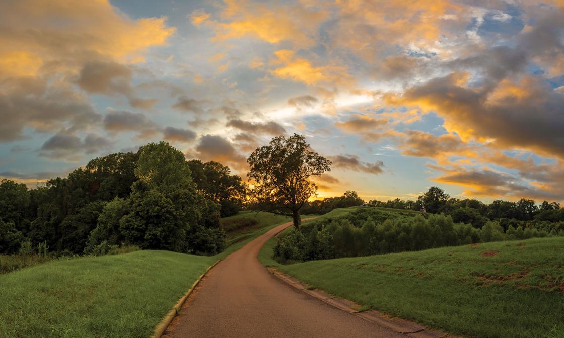 A paved path leads up a hill to a monument in the far distance, under a colorful sunset.