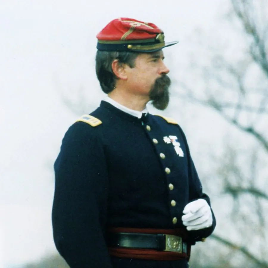Man with dark hair and a mustache wearing a Civil War-era military uniform and red kepi-style cap.