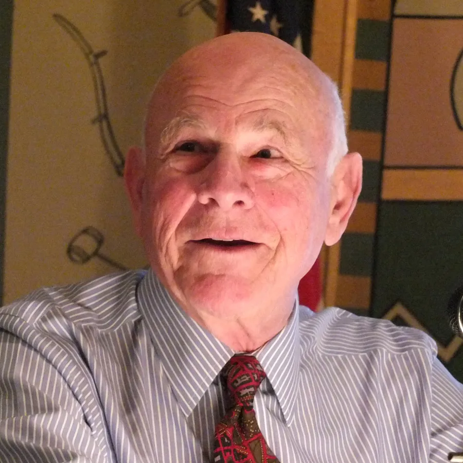 Older man wearing a striped dress shirt and patterned tie, with a colorful background behind him.