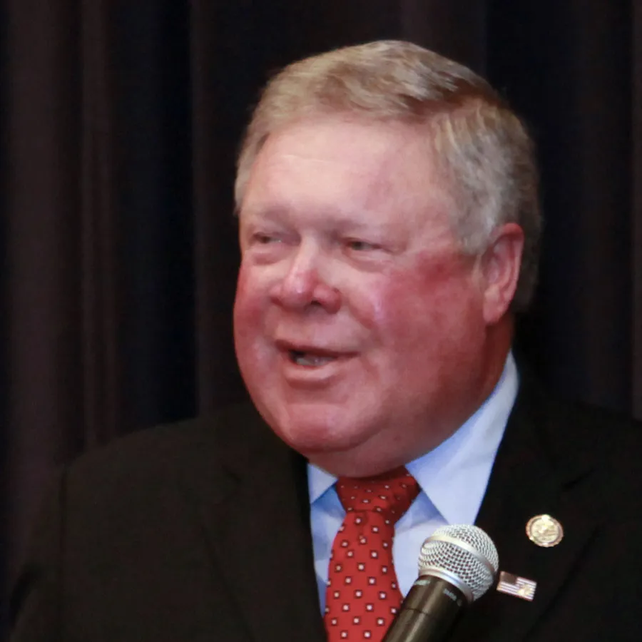 Man speaking into a microphone, wearing a dark suit, light blue shirt, and red patterned tie.