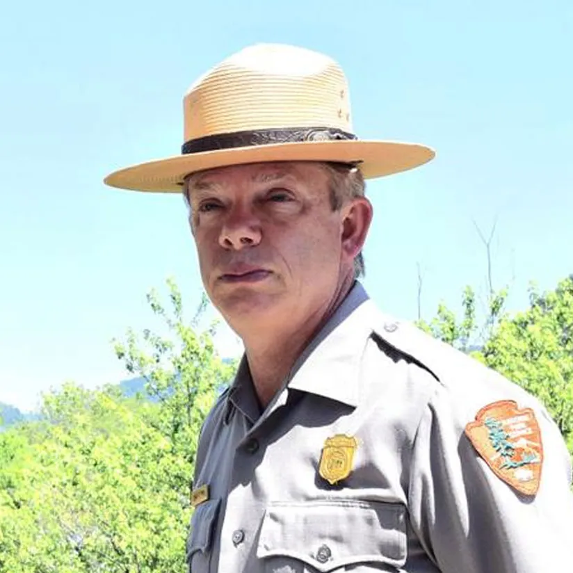 Man wearing a National Park Service uniform and broad-brimmed ranger hat, outdoors against trees and a blue sky.