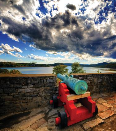 Bright colorful photo showing a Revolutionary War cannon with a red base overlooking water with puffy clouds filling the sky